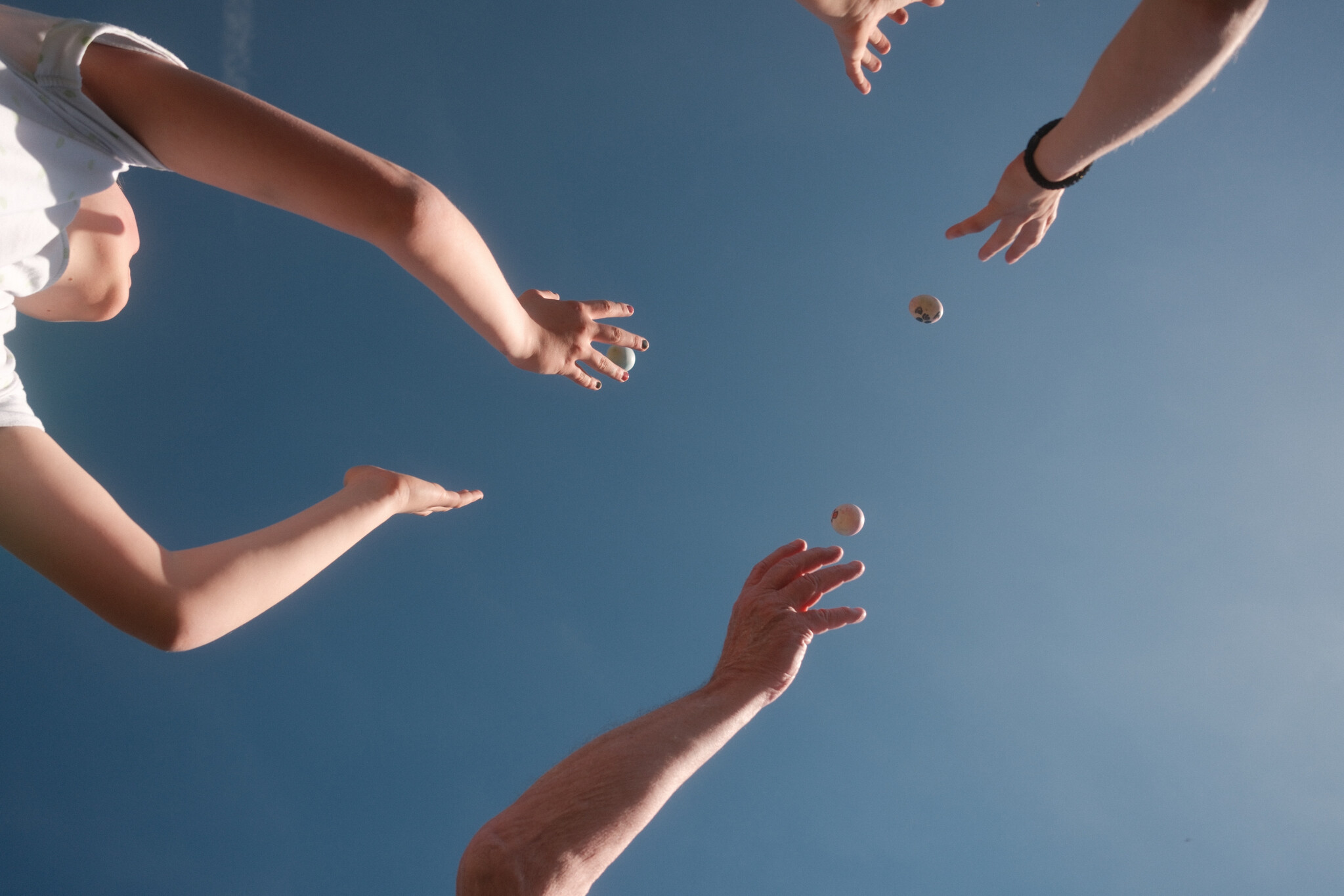 Low-angle view of three people's arms and hands reaching up toward the sky, throwing and catching Easter eggs. A child in a white t-shirt is visible on the left. Two eggs are mid-air at the center of the image. The background is a clear, uniform blue sky.