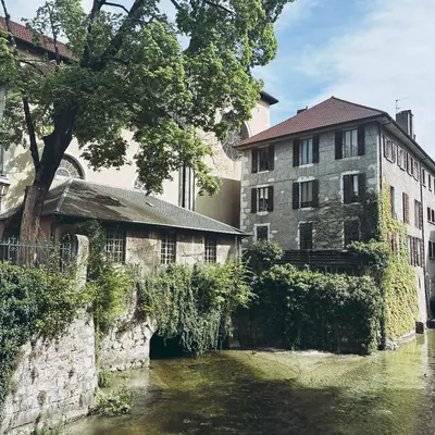 A tranquil setting in the old town of Annecy, showcasing a stone building covered in ivy beside a peaceful canal, with a large tree providing shade.