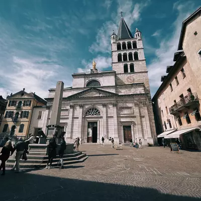 A view of a historic church in the old town of Annecy, with a prominent clock tower and an open square in front. People are walking around, enjoying the sunny day.