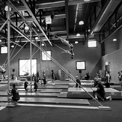 The Wettingen gymnastics club presents a swinging rings performance in a gymnasium. Several gymnasts execute acrobatics while coaches and spectators observe. The scene is in black and white, with visible metal architecture and indoor lighting.