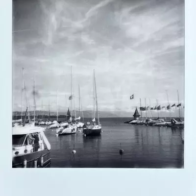 Black and white Polaroid photograph of Morges harbor on Lake Geneva, showing several sailboats moored at the marina with their masts and rigging visible, flags flying, and a cloudy sky overhead.