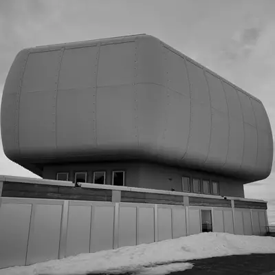 A modern building with a smooth and rounded rectangular design on top of a platform. The structure is set against a cloudy sky with patches of snow on the ground.