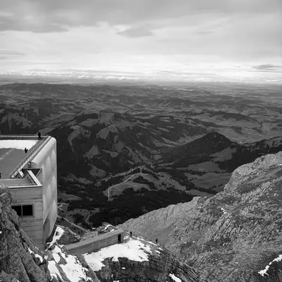 A black and white view from Säntis, showing a modern building perched on a rocky mountain ledge. The structure overlooks a vast, rolling landscape with scattered trees and fields, beneath a cloudy sky.