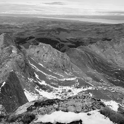 A breathtaking view from the Säntis mountain, overlooking rugged peaks with patches of snow. The landscape extends to a distant lake and sprawling countryside under a cloudy sky.