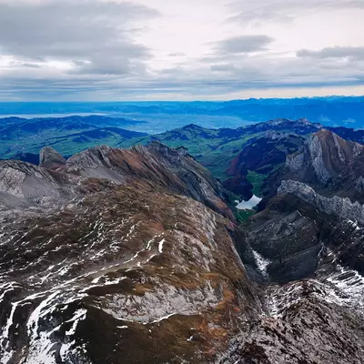 A panoramic view from Säntis, showing a rugged mountain range with patches of snow and a lush green landscape in the distance. The sky is overcast, giving a dramatic backdrop to the scene.