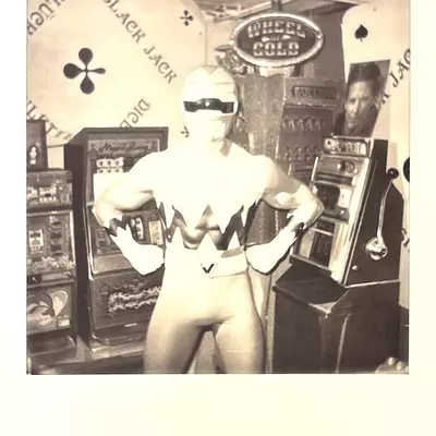 Black and white Polaroid of a mannequin doll wearing a superhero-style costume with zigzag pattern, displayed in a vintage arcade setting with 'Wheel of Gold' signage and slot machine props at Dreamfactory museum in Degersheim
