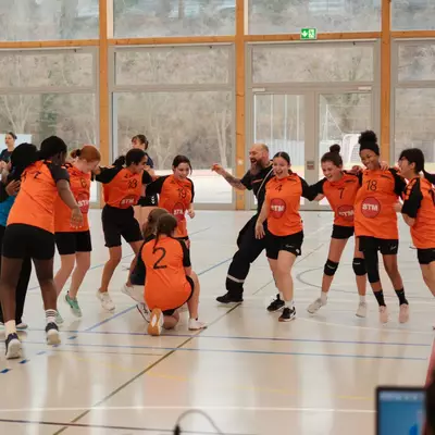 L'équipe du HBC Moudon en maillot orange célèbre ensemble sur le terrain, joueuses et entraîneur se réunissant dans une explosion de joie