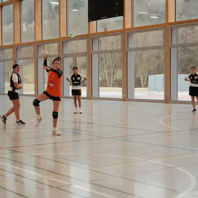 Joueuse du HBC Moudon en maillot orange s'apprêtant à effectuer un tir, face à des défenseuses du HC Vevey en maillot noir et blanc, dans une salle de sport aux grandes baies vitrées