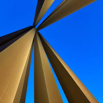 Golden converging lines meeting at a focal point against an intense blue sky. The metallic bands create a dynamic radial pattern, with their sharp edges cutting through space, forming a striking geometric composition.