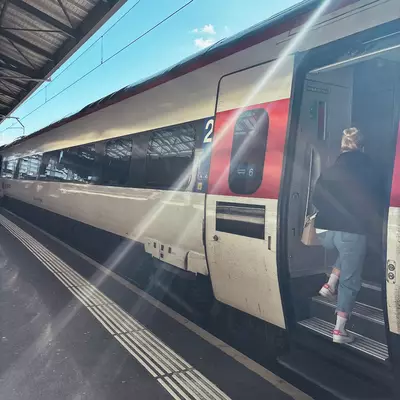 Train at platform with pronounced horizontal lines on its bodywork. The tactile guide strips on the ground create parallel lines, while overhead power lines draw linear patterns against the blue sky. The carriage features a design with red and white decorative stripes, creating a layered linear composition.