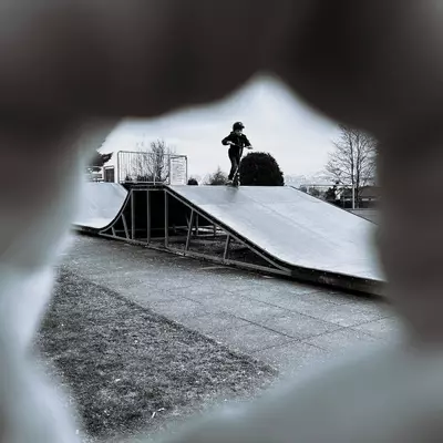 View framed through a blurry, irregular opening, capturing a skater in black at the top of a skate ramp. The natural framing creates a striking contrast between the dark areas in the foreground and the skater's silhouette against the bright sky. The black and white composition enhances the dramatic effect of the scene.