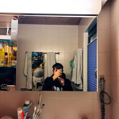 Self-portrait in a bathroom mirror framed by beige tiles. Everyday objects - toothbrushes, towels, and accessories - line the composition, while a Ninjago decorative towel adds a pop of color in the background. The rectangular frame of the mirror naturally structures the image.