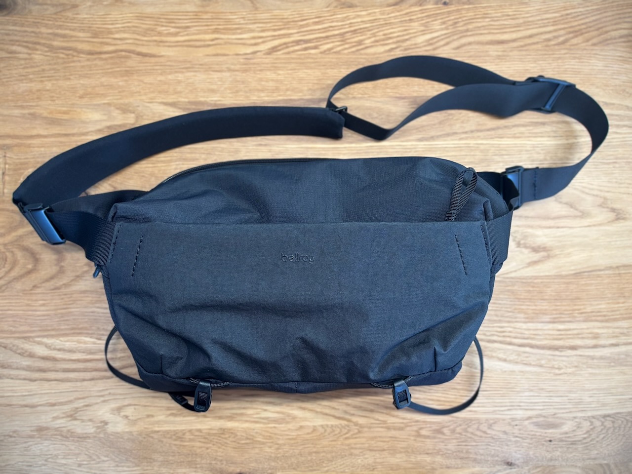 Front view of a closed Bellroy Venture Camera Mag in black, laid flat, showing the minimalist design, the Bellroy logo, the front zipper pocket, and the shoulder strap.