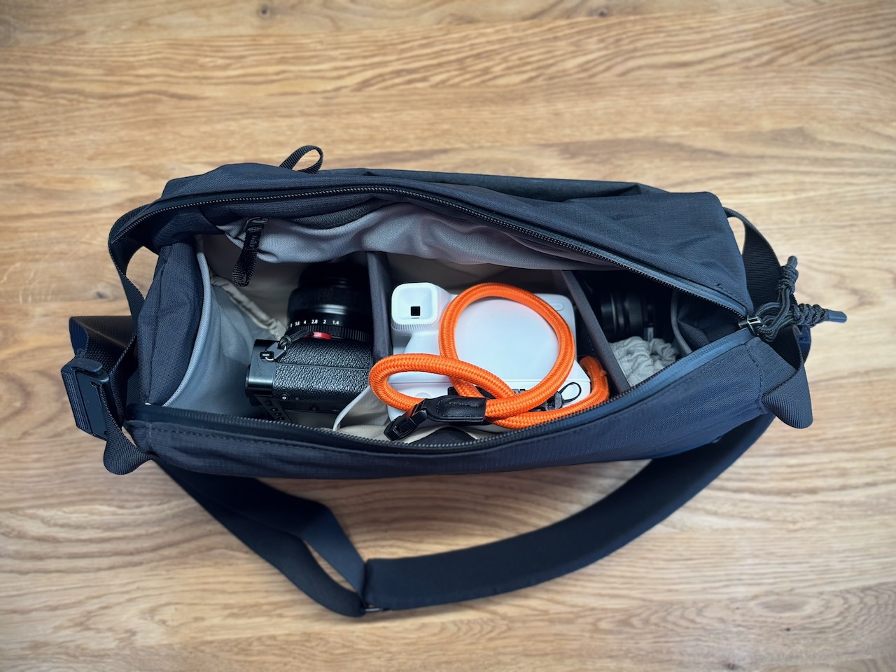 Top view of an open Bellroy Venture Camera Mag showing a Fujifilm camera with lens, a Polaroid Flip instant camera with an orange wrist strap, and a second lens, organized in three compartments with fixed dividers.