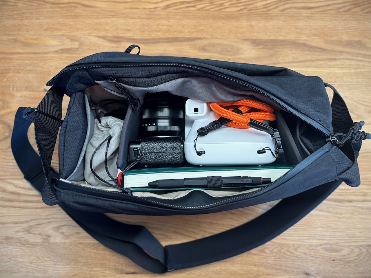 Top view of an open Bellroy Venture Camera Mag containing a Fujifilm camera, a white Polaroid Flip camera with an orange strap, a cleaning cloth, and an A5 notebook with a pen resting on top.