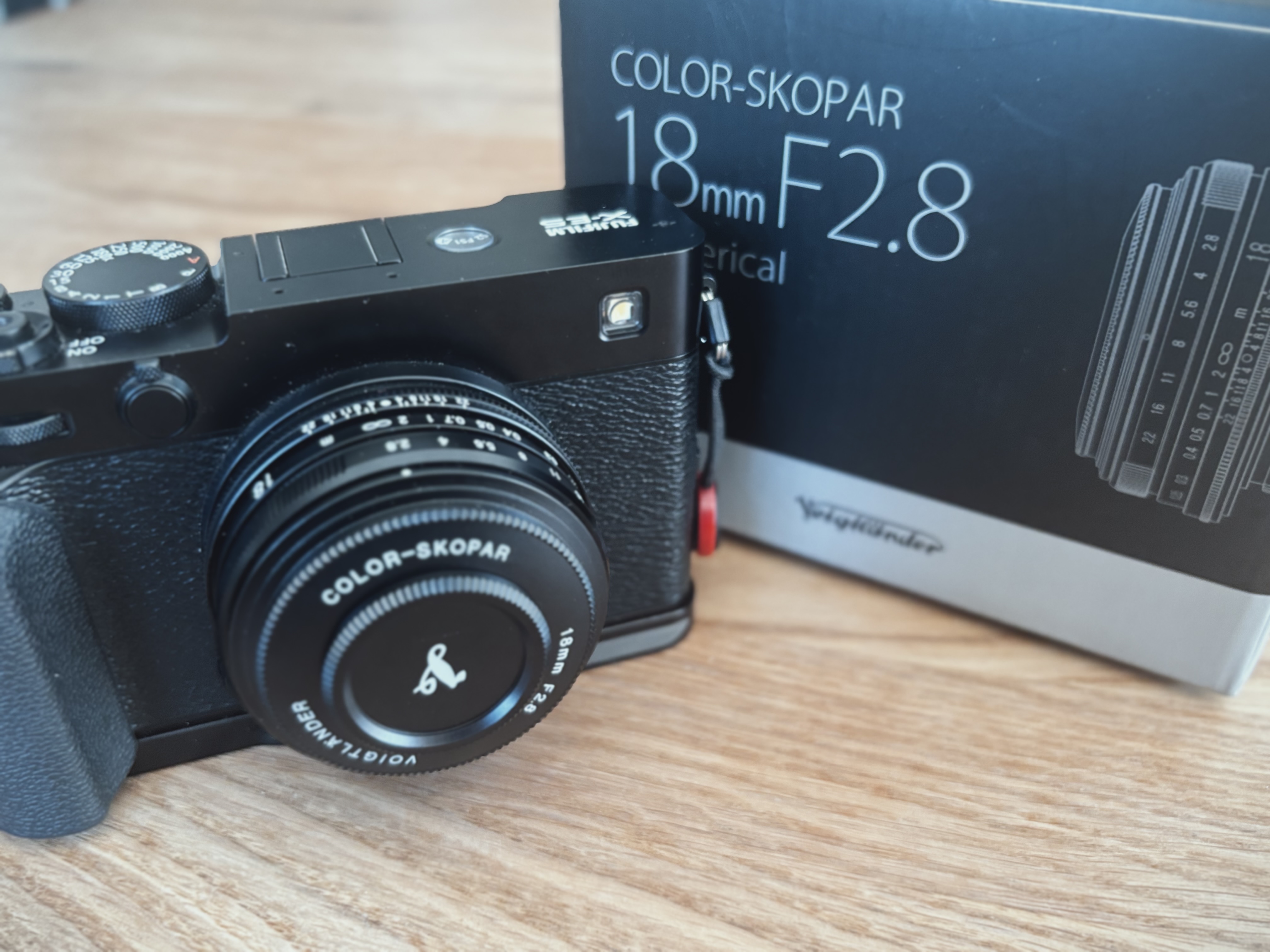 Fuji X-E5 camera fitted with a Voigtländer Color-Skopar 18mm F2.8 lens, next to its retail box on a wooden surface.