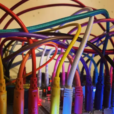 Colorful patch cables in red, orange, yellow, green, blue, and purple are tangled together, connected to various modules in a modular synthesizer setup. The cables create an intricate web of connections against a warm-toned background, showcasing the complex wiring typical of modular synth patching.