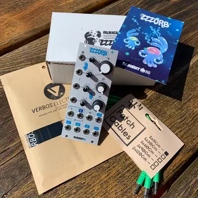 A collection of synthesizer modules and documentation laid out on a wooden surface, including a white Verbos Electronics module with multiple knobs and controls, a blue Pizzorb module box, kraft paper packaging labeled 