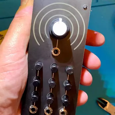 A hand holds a dark gray synthesizer module with a white illuminated knob at the top surrounded by concentric circles, a gold input jack below it, and a grid of nine smaller knobs arranged in three rows at the bottom against a turquoise workbench.