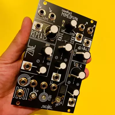 A hand holding a black circuit board synthesizer module with white knobs, gold jacks, and handwritten labels against a bright yellow background. The module appears to be the Mimeophon, a compact electronic music device with multiple controls and connections arranged in a grid pattern.