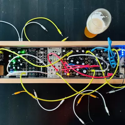 A DIY Eurorack synthesizer case with wooden sides containing two rows of electronic modules with colorful wiring in yellow, green, blue, and red. A glass of beer sits in the upper right corner of the dark surface. Multiple cables and connectors extend from the modules.