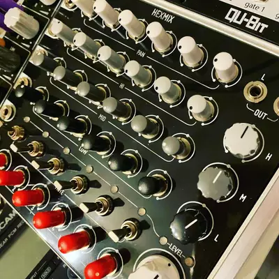 Close-up of a black audio mixing console with multiple white rotary knobs arranged in vertical columns, red indicator lights on the left side, and gold-plated connectors. The device is labeled 