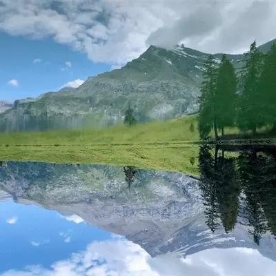 A pristine alpine lake mirrors snow-capped mountains, dense evergreen forests, and a bright blue sky dotted with white clouds. Lush green meadows stretch along the shoreline, creating a serene mountain landscape. The water's glassy surface reflects the surrounding peaks and vegetation in perfect clarity.