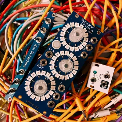 A blue electronic synthesizer module with three rotary knobs sits among a tangle of colorful audio cables in red, yellow, and blue. The module has white labels and circular dials marked with positions around each knob. A small white control panel with input/output jacks is visible on the right side of the arrangement.