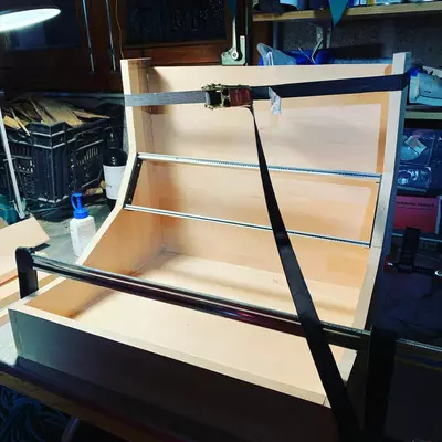 A DIY Eurorack case under construction with light wooden shelving, metal rails, and black support straps, displayed on a workbench surrounded by electronic equipment and tools.