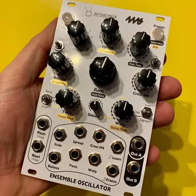 A Matthias Puech Ensemble Oscillator Eurorack module held in someone's hand against a yellow background. The white faceplate features multiple black knobs for controlling parameters like Frequency, Timbre, Detune, and various modulation options, with labeled sections for Root, Cross FM, and outputs A and B at the bottom.