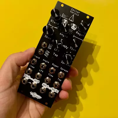 A hand holds a black Eurorack synthesizer module against a bright yellow background. The module features numerous small knobs, jacks, and switches with white text labels for various parameters including filters, oscillators, and modulation controls. The compact device appears to be a complex audio processing or synthesis tool designed for modular synthesizer systems.