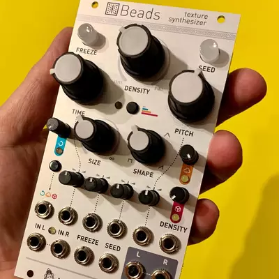 A hand holding a Beads texture synthesizer module against a bright yellow background. The cream-colored Eurorack module features multiple black knobs and small buttons labeled with parameters like Freeze, Density, Time, Seed, Pitch, Size, and Shape, along with input/output jacks at the bottom.