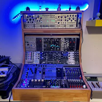A wooden DIY Eurorack case with multiple rows of modular synthesizer modules illuminated by blue LED lighting. The case contains various patch cables, knobs, and control panels across four separate shelves, with additional modules visible on top. The setup is photographed in a studio environment with blue ambient lighting in the background.