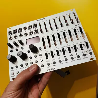 A hand holds a white Metropolix synthesizer against a yellow background, displaying its compact control layout with multiple knobs, faders, buttons, and a small screen for electronic music production.