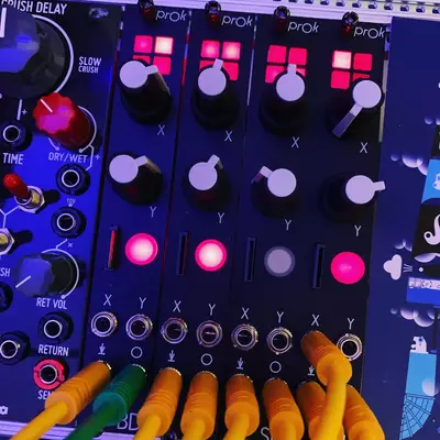 A modular synthesizer setup with multiple ProK units displaying illuminated pink and magenta buttons and controls arranged in a grid pattern. Orange and green patch cables are connected to the bottom of the units. The interface includes X and Y labeled controls with corresponding knobs and illuminated indicator lights throughout the blue-tinted faceplate.