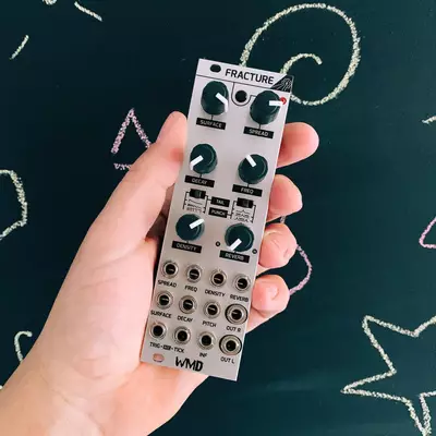 A hand holds a white WMD Fracture Eurorack module against a dark teal background with hand-drawn circular designs. The module displays six vertical channels with knobs, buttons, and input/output jacks for audio processing. A coiled white cable is attached to the top of the module.