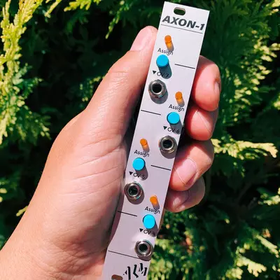 A hand holds a white Eurorack module labeled AXON-1 against a blurred green foliage background. The module displays four identical sections, each with an orange knob, blue button, and silver jack socket, with 