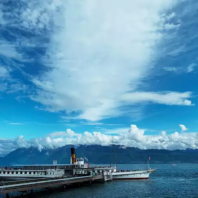 A vintage paddle steamer with red and black smokestacks is docked at a pier on a large alpine lake, with snow-capped mountains visible across the water under a partly cloudy blue sky.