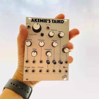 A person holding a beige Akemie's Taiko synthesizer module with multiple knobs and controls arranged in rows, including labeled parameters for Fmod Start, Fmod End, Pitch, Release, Algo, Trigger, and other synthesis controls.