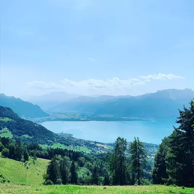 A sweeping alpine valley view with turquoise-blue lake surrounded by forested mountainsides and distant peaks under a partly cloudy sky.