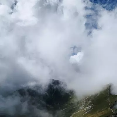 Dense clouds engulf towering mountain peaks with steep rocky slopes, obscuring a valley below where green vegetation clings to the hillsides in this dramatic alpine landscape.