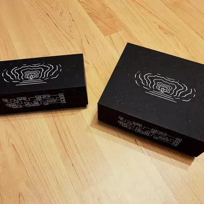 Two black rectangular boxes with white line-art designs on their lids sit on a light wood floor. Both boxes feature similar concentric circular patterns with geometric elements. Small white text is visible on the sides of each box.