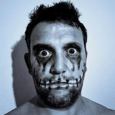A man with dramatic black makeup applied around his eyes and mouth, creating dark streaks down his face, stares directly at the camera with an intense expression against a plain light blue background.