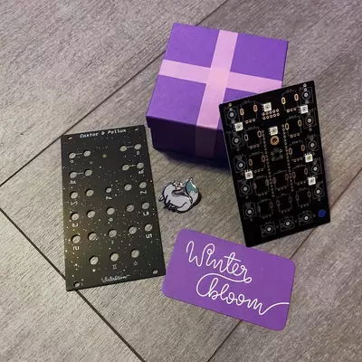 A purple gift box with a cross on top sits among two black synthesizer keyboards, a silver ring, and purple cards labeled 