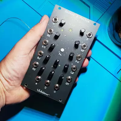 A hand holds a black audio mixer with multiple knobs and faders against a bright blue surface, with cables and equipment visible in the background.