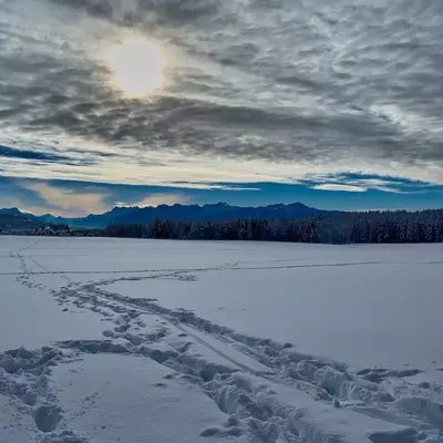 A frozen lake stretches across the foreground with tire tracks and footprints marking the snow, while forested shorelines and snow-capped mountains line the horizon under a dramatic cloudy sky with breaks of blue light.