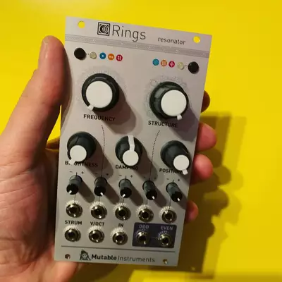A hand holds a Mutable Instruments Rings resonator module against a bright yellow background. The gray module features two large knobs labeled Frequency and Structure at the top, with smaller knobs below for B. Offset, Damping, and Position, plus additional controls for Strum, Voct, In, and Odd/Even parameters.