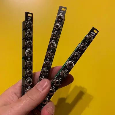 A hand holding three vertical black modular synthesizer modules against a yellow background. Two modules labeled 