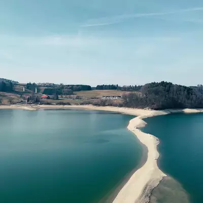 A turquoise alpine lake with a distinctive curved sandy sandbar dividing the water, surrounded by forested hillsides and scattered buildings nestled along the shoreline under a clear blue sky.