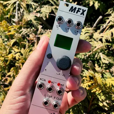 A white modular synthesizer module labeled MFX is held in someone's hand against a blurred green foliage background. The compact device features a small LCD screen, three knobs at the top, a large black central knob, and multiple input/output jacks arranged in rows below.